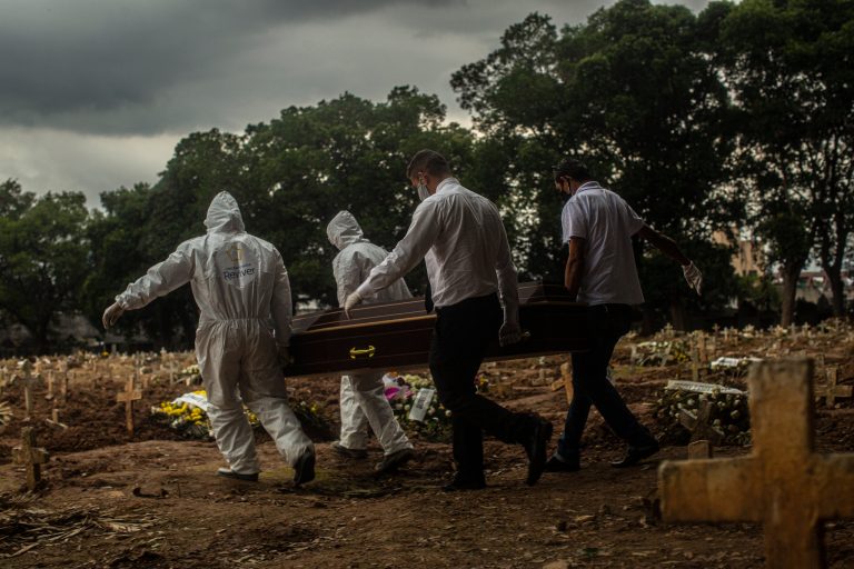 Workers in white protective suits carry a coffin through a cemetery in Rio de Janeiro under a gray sky. Credit: Dado Galdieri for The New York Times.