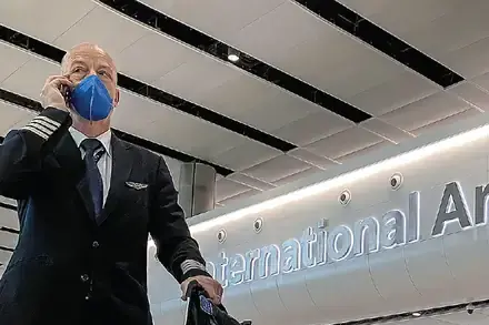A masked airline pilot and masked travelers beneath an International Arrivals sign. Credit: Toby Melville/Reuters.